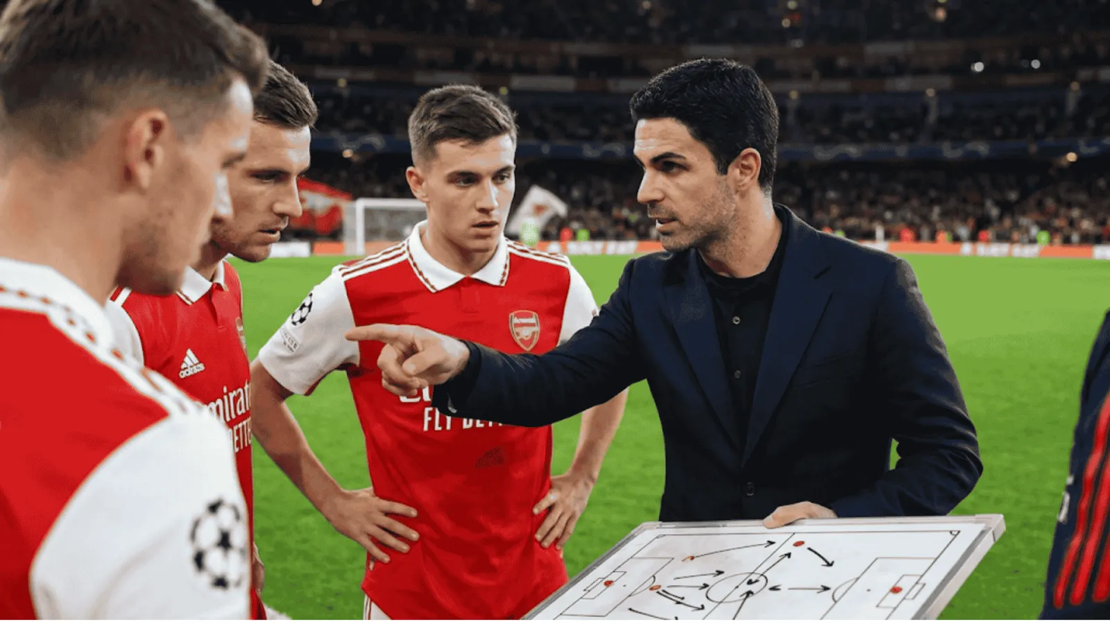 A photorealistic, intimate close-up shot from the sideline showing Arsenal manager Mikel Arteta gesturing and pointing toward a tactical whiteboard. He is surrounded by focused Arsenal players in their red and white kits
