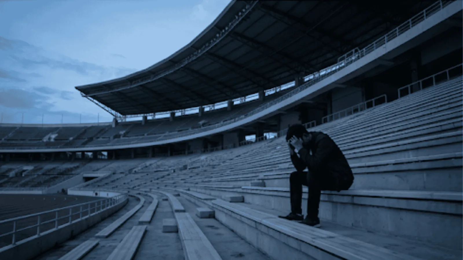 Candid wide-angle of a solitary defeated figure sitting on empty stadium bleachers at dusk under atmospheric floodlight shadows.